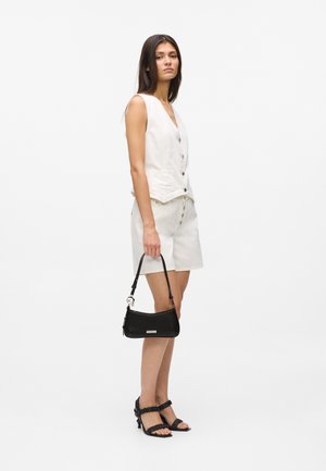 Fiatal nő fehér ujjatlan, gombos mellényben és rövidnadrágban, fekete táskával a kezében, fekete magassarkú szandált viselve, oldalra fordulva áll.