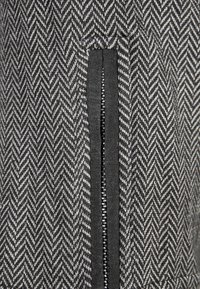 Kostkovaná látka v černobílé barvě obsahuje zipový detail, má texturovaný tkaný vzor a solidní černé akcenty podél okrajů.