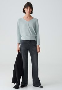 Helle blaue Rippstrickpullover mit V-Ausschnitt und langen Ärmeln, kombiniert mit schwarzen Schlagjeans. Eine schwarze Jacke wird in der linken Hand gehalten.