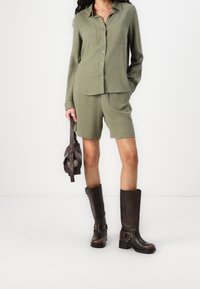 Chemise vert olive avec deux poches avant, design à boutons, assortie à un short coordonné. Chaque jambe est coupée au-dessus du genou, portée avec des bottes marron montant jusqu'aux genoux.