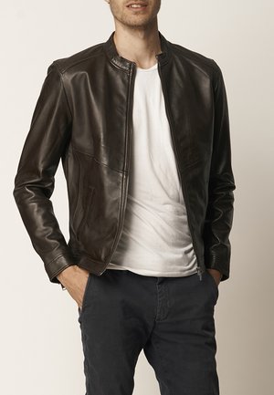 Man in een donkerbruin leren jas over een wit T-shirt en donkere broek, staand met handen in de zakken tegen een effen achtergrond.