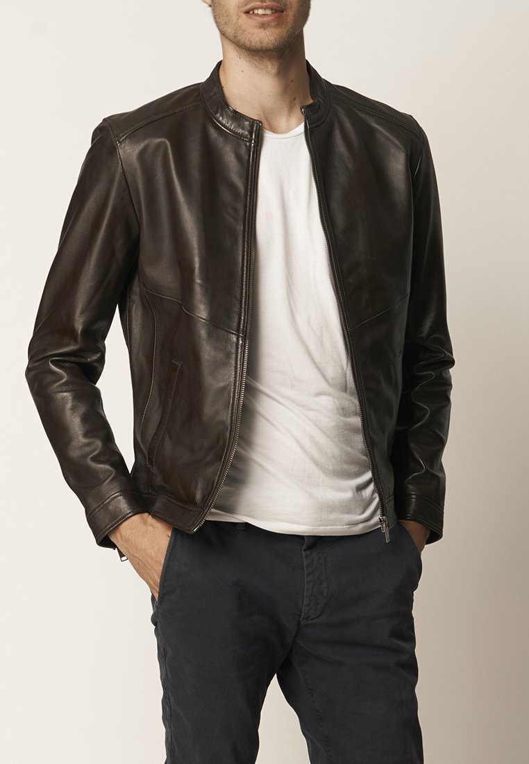 Hombre vestido con una chaqueta de cuero marrón oscuro sobre una camiseta blanca y pantalones oscuros, de pie con las manos en los bolsillos contra un fondo liso.