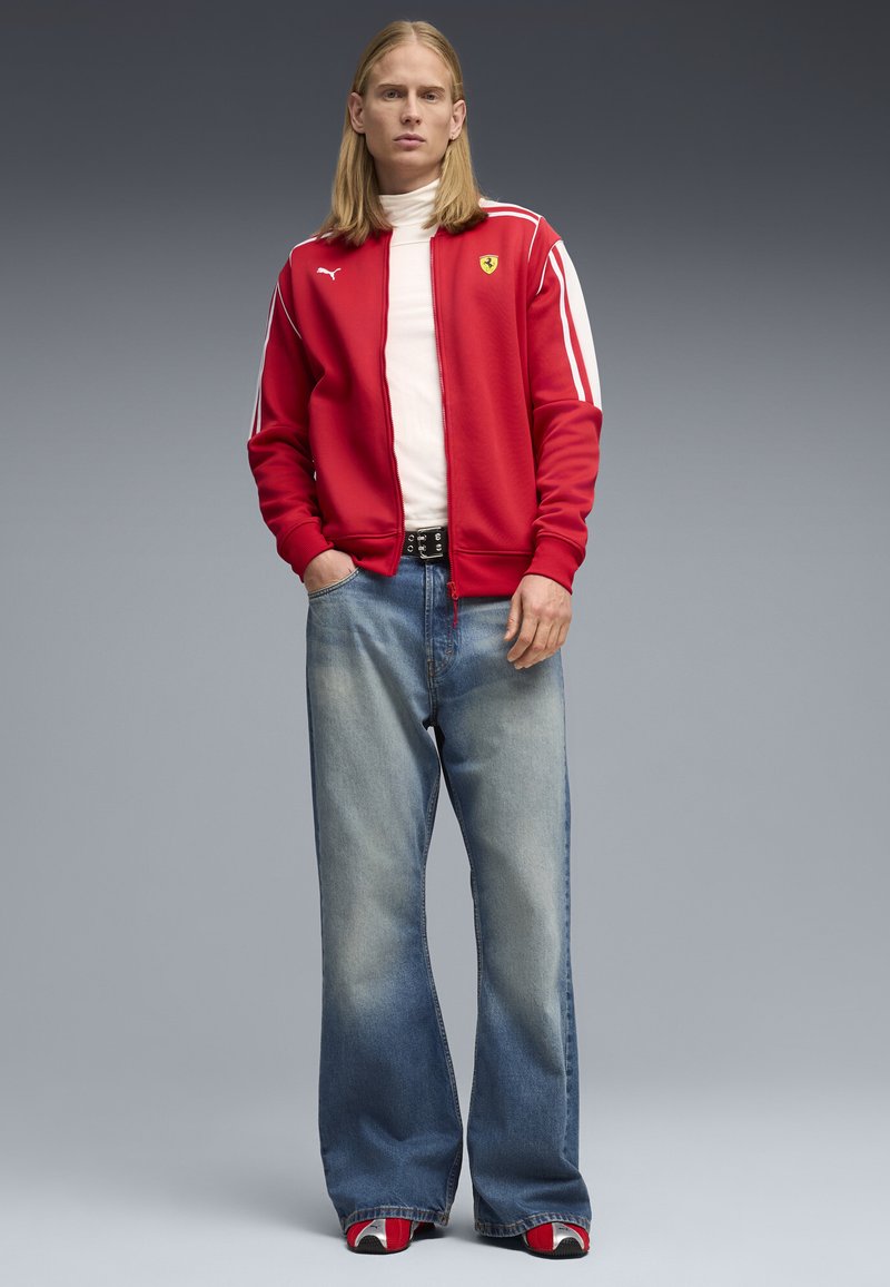 Jeune personne aux longs cheveux blonds portant une veste rouge Ferrari Puma, un col roulé blanc, un jean large délavé et des baskets rouges.