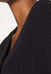 Primer plano del cuello y hombro de una persona que lleva un suéter de punto texturizado negro con un escote en V profundo.