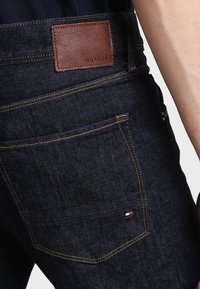 El bolsillo trasero de unos jeans azul oscuro con costuras de contraste, una pequeña bandera roja, blanca y azul de Tommy Hilfiger, y un parche de cuero marrón de la marca Hilfiger.