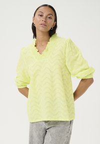 Blusa gialla con motivo a zig zag, colletto arricciato, maniche a sbuffo e tessuto testurizzato. Caratterizzata da una vestibilità comoda e materiale leggero.