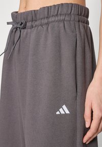 Sive bombažne trenerke z elastičnim pasom in vrvico, ki vključujejo stranske žepe in belo Adidasovo logo blizu roba.