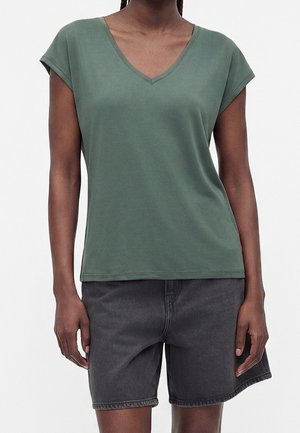 Persoon draagt een groen T-shirt met korte mouwen en V-hals en donkergrijze denim shorts, staand met armen ontspannen langs het lichaam.