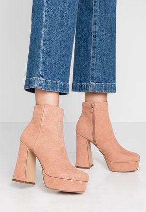 Pieds chaussés de bottines à plateforme en suède pêche avec des talons épais, associées à un jean bleu raccourci sur un fond uni.