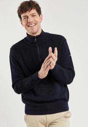 Marineblauer Strickpullover mit hohem Kragen und kurzer Reißverschlussöffnung. Rippstruktur an Ärmeln und Saum. Das Modell klatscht in die Hände und trägt beiges Hosen.