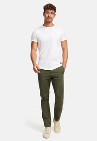 Camiseta blanca de manga corta combinada con pantalones verdes ajustados y zapatillas beige en un modelo frente a un fondo neutro.