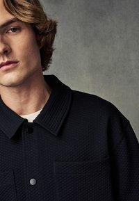 Schwarze steppjacke mit breitem Kragen, Fronttasche und Knopfverschluss. Der Stoff hat ein strukturiertes Aussehen und eine glatte, matte Oberfläche.