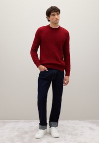 Roter gestrickter Pullover mit strukturiertem Rautenmuster, kombiniert mit dunkelblauen Jeans und weißen Turnschuhen. Der Pullover hat einen Rundhalsausschnitt und gerippte Bündchen.