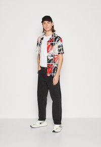 Camisa de manga corta en negro, rojo, blanco y beige con patrones gráficos; combinada con jeans negros y zapatillas blancas. Lleva una gorra negra.