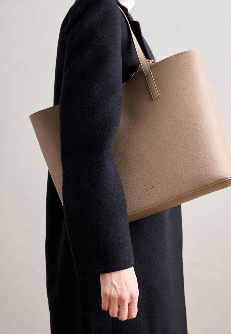 Personne en manteau noir portant un grand sac cabas en cuir beige sur l'épaule, devant un fond clair uni.
