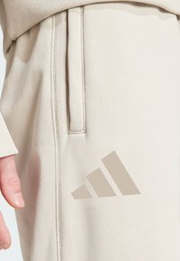 Beige athletic joggers made of soft fabric, featuring a side pocket and subtle tonal Adidas logo in the lower right. Textured finish.