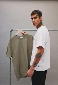 Hombre con tatuajes en el brazo y el cuello sosteniendo una camiseta verde oliva en una percha en un entorno de estudio neutro.