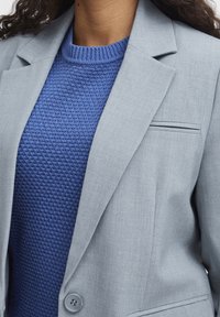 Světle šedý sak se strukturovaným designem a zapínáním na jeden knoflík, v kombinaci s texturovaným modrým svetrem. Obsahuje náprsní kapsu s tenkým akcentem.