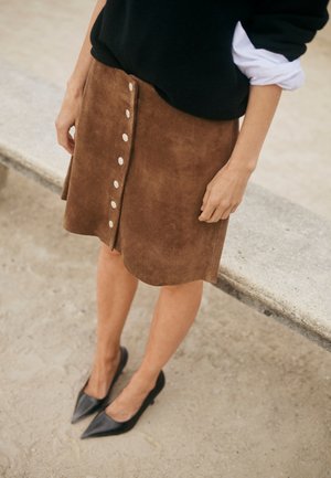 Persona che indossa una gonna in camoscio marrone con bottoni sul davanti, ballerine nere con punta appuntita, maglione nero con maniche della camicia bianca arrotolate.