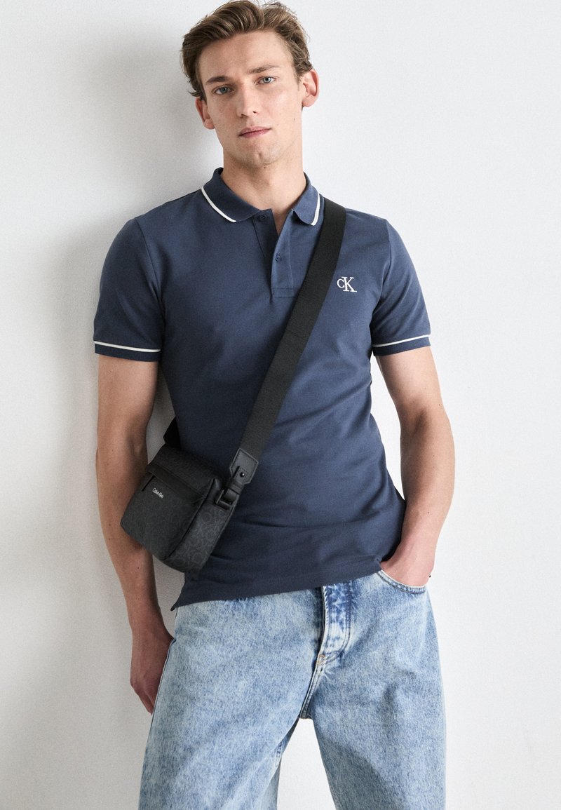 Marineblaues Poloshirt mit weißen Akzenten und CK-Logo. Kombiniert mit hellblauen Jeans aus Denim. Schwarze Umhängetasche mit texturiertem Design.