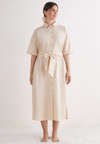 Beiges Leinenhemdkleid mit kurzen Ärmeln, Knopfleiste und einem selbstgebundenen Gürtel in der Taille. Mit seitlichen Schlitzen für mehr Bewegungsfreiheit.