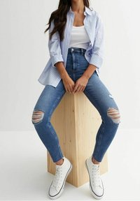 Mujer con jeans skinny azul rasgados, zapatillas blancas, camiseta sin mangas blanca y camisa de botón a rayas azul claro sentada sobre un bloque de madera.