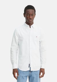 Camicia bianca con bottoni, caratterizzata da una sottile trama a righe, maniche lunghe e un piccolo logo. Abbinata a jeans blu.