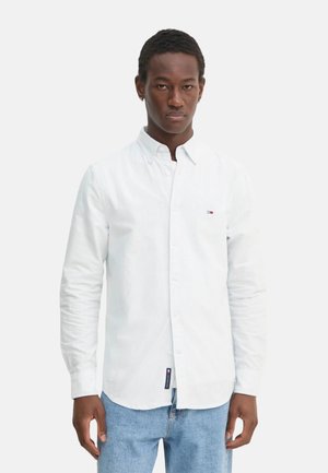 Camisa blanca abotonada con una sutil textura a rayas, mangas largas y un pequeño logotipo. Combinada con jeans azules.