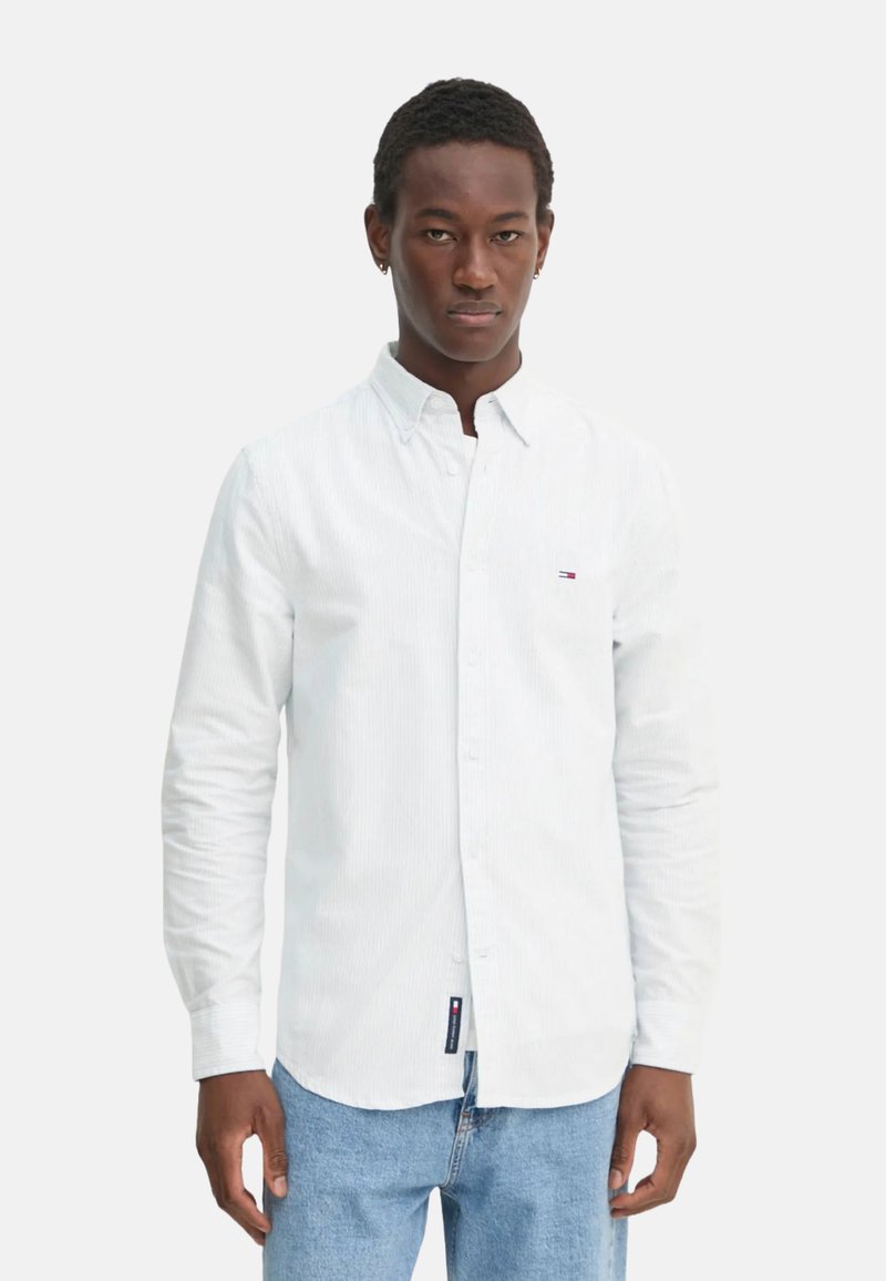 Camicia bianca con bottoni, caratterizzata da una sottile trama a righe, maniche lunghe e un piccolo logo. Abbinata a jeans blu.