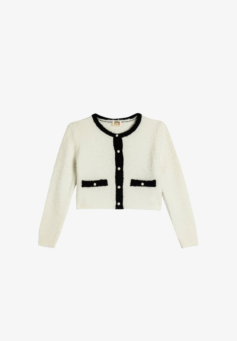 Weißer, kürzer geschnittener Cardigan mit einer flauschigen Textur, schwarzer Kante und zwei schwarzen Taschen. Verfügt über perlenähnliche Knöpfe auf der Vorderseite.