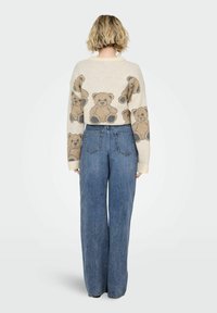 Przycięty beżowy sweter z wzorem misia; zestawiony z szerokimi niebieskimi dżinsami. Sweter ma ribbowe mankiety i dół.