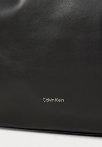 Borsa nera in pelle con una texture liscia e un design minimalista. Il logo dorato "Calvin Klein" vicino alla parte inferiore aggiunge un branding sottile.