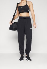 Svart sportklädsel med en kort sport-BH med tre vita ränder och svarta sweatpants med dragsko, kombinerat med svarta sneakers och en väska.