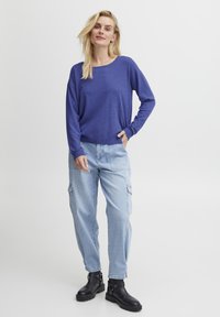 Pulover lung mov cu mânecă lungă, asortat cu pantaloni cargo de culoare albastră deschisă și cizme negre până la gleznă. Puloverul are o croială lejeră și decolteu rotund.