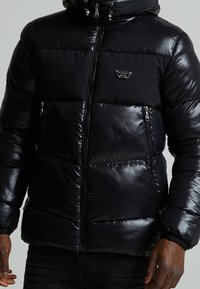 Schwarze Puffjacke mit glänzender Oberfläche, Reißverschluss vorne, zwei seitlichen Taschen und einem Logo-Patch auf der linken Brust. Mit Kapuze und elastischen Bündchen.