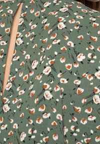 Zielona tkanina w kwiatowy wzór z małymi białymi i brązowymi kwiatami, z dekoltem w kształcie łezki i miękką, teksturowaną powierzchnią.