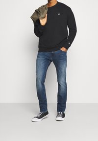 Sudadera de algodón negra con cuello redondo, combinada con jeans de mezclilla azules. Se llevan zapatillas de lona en blanco y negro. Se sostiene una chaqueta verde oliva.