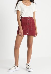 Jupe en jean rouge avec des boutons sur le devant, associée à un t-shirt léger à col en V, et des baskets blanches avec des pois noirs. Texture lisse et design décontracté.