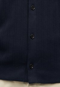 Κοντινή λήψη ενός μπλε σκούρου ριμπ πλεκτού ζακέτα με μαύρα κουμπιά, που φοριέται πάνω από μπεζ παντελόνι.