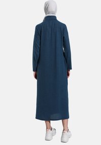 Abito camicia lungo di colore blu scuro, con colletto, maniche lunghe e vestibilità comoda, realizzato in un tessuto testurizzato. Indossato con sneakers bianche e voluminose.