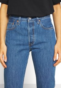 Jeans de mezclilla azul de tiro alto con un acabado desgastado, diseño de cinco bolsillos y herrajes en tono cobre, combinados con una blusa negra.