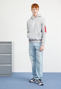 Felpa grigia con logo frontale e tasca con zip rossa. Indossata con jeans azzurri e sneakers bianche e nere. Tessuto morbido e casual.