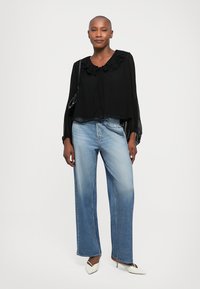 Vrouw staan in een zwarte ruches blouse, lichtblauwe wijde jeans, witte puntige hakken en een zwarte schoudertas.