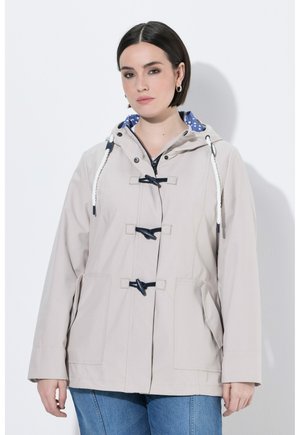 Vrouw die een beige gewatteerde jas met capuchon draagt, voorzien van koordsluitingen, witte trekkoorden, een blauwe voering met witte polkadots en een blauwe spijkerbroek, staand tegen een witte achtergrond.