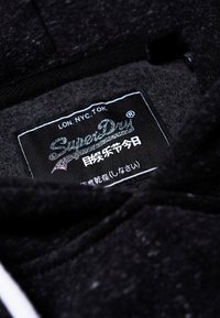 Etiqueta de la prenda en color negro que muestra la marca "SuperDry", abreviaturas de ciudades (LON, NYC, TOK) y caracteres japoneses.