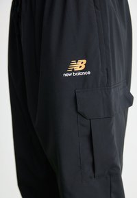 Black New Balance pants with cargo pocket on side and gold logo above pocket on thigh.