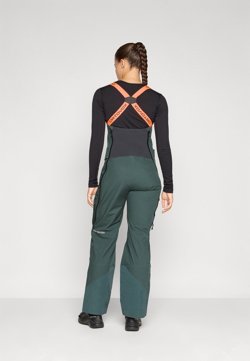 Grüne Skihosen aus schwarzem Softshell-Material mit orangefarbenen Trägern. Mit Reißverschlüssen an den Seiten und einem taillierten Design, geeignet für den Einsatz im Freien.