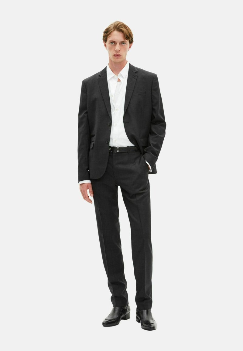 Donkergrijs pak met een gestructureerde blazer en op maat gemaakte broeken. Witte shirt eronder. Zwarte riem en glanzende zwarte schoenen. Eenvoudig, modern ontwerp.