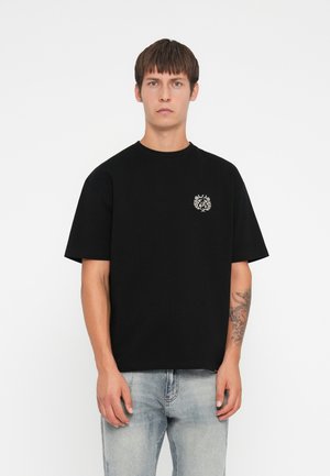 T-shirt en coton noir avec col ras du cou et manches courtes, arborant un emblème décoratif blanc sur le côté gauche de la poitrine. Porté avec un jean en denim clair.