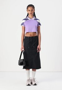 Top atlético roxo curto com mangas em blocos de cores, saia preta com cordão e mala preta de ombro. A modelo usa ténis claros e meias brancas.